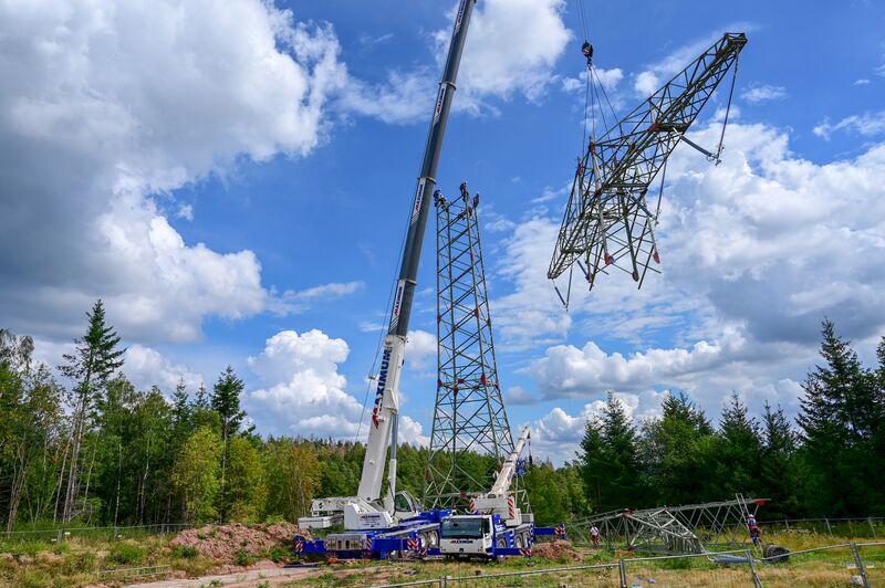 Ein großer Mobilkran richtet auf einer Freifläche am Waldrand einen Gittermast einer Hochspannungsfreileitung auf. Mehrere Monteure begleiten die Montage, während weitere Mastsegmente am Boden bereitliegen. Über der Baustelle erstreckt sich ein blauer Himmel mit großen weißen Wolken.