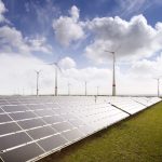 Großer Solarpark mit langen Reihen von Photovoltaikmodulen vor mehreren Windenergieanlagen. Die Anlage erstreckt sich über eine offene Landschaft unter einem wolkigen Himmel und veranschaulicht die kombinierte Nutzung von Solar- und Windenergie zur Stromerzeugung.