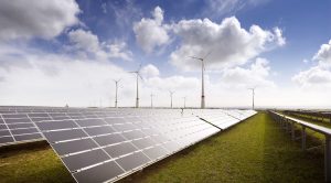 Großer Solarpark mit langen Reihen von Photovoltaikmodulen vor mehreren Windenergieanlagen. Die Anlage erstreckt sich über eine offene Landschaft unter einem wolkigen Himmel und veranschaulicht die kombinierte Nutzung von Solar- und Windenergie zur Stromerzeugung.