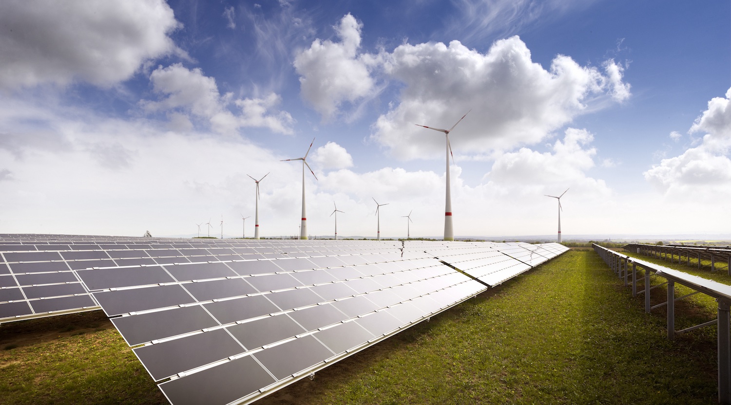 Großer Solarpark mit langen Reihen von Photovoltaikmodulen vor mehreren Windenergieanlagen. Die Anlage erstreckt sich über eine offene Landschaft unter einem wolkigen Himmel und veranschaulicht die kombinierte Nutzung von Solar- und Windenergie zur Stromerzeugung.
