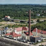 Luftaufnahme der Energiezentrale Wedau mit historischem Backsteinschornstein und sanierten Gebäuden. Im Vordergrund sind die neue Energiezentrale sowie angrenzende Gebäude des Quartiers zu sehen. Im Hintergrund erstrecken sich Waldflächen, eine Straßenbrücke und weitere Bebauung. Die Aufnahme zeigt die Einbindung der Anlage in das neu entstehende Stadtquartier Wedau.