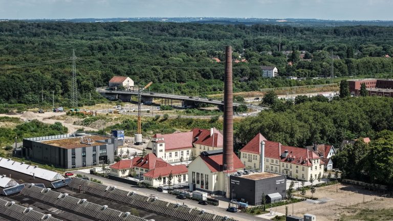 Luftaufnahme der Energiezentrale Wedau mit historischem Backsteinschornstein und sanierten Gebäuden. Im Vordergrund sind die neue Energiezentrale sowie angrenzende Gebäude des Quartiers zu sehen. Im Hintergrund erstrecken sich Waldflächen, eine Straßenbrücke und weitere Bebauung. Die Aufnahme zeigt die Einbindung der Anlage in das neu entstehende Stadtquartier Wedau.