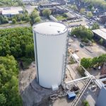 Luftaufnahme eines zylindrischen Wärmespeichers auf dem Gelände eines Heizwerks in Gelsenkirchen. Der weiß verkleidete Speicher steht neben Industriegebäuden, einem Schornstein und Rohrleitungen. Rundherum sind Straßen, Parkplätze, Gewerbebauten und Grünflächen zu sehen. Der Wärmespeicher ist in das Fernwärmenetz des Standorts eingebunden.
