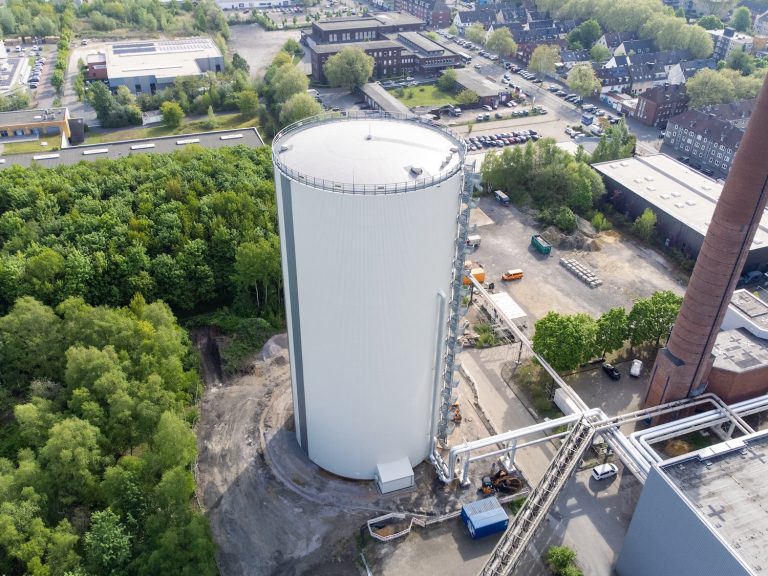 Luftaufnahme eines zylindrischen Wärmespeichers auf dem Gelände eines Heizwerks in Gelsenkirchen. Der weiß verkleidete Speicher steht neben Industriegebäuden, einem Schornstein und Rohrleitungen. Rundherum sind Straßen, Parkplätze, Gewerbebauten und Grünflächen zu sehen. Der Wärmespeicher ist in das Fernwärmenetz des Standorts eingebunden.