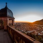 Blick von der Terrasse des Heidelberger Schlosses auf die Altstadt bei Sonnenuntergang. Im Vordergrund sind die historische Schlossmauer, eine Balustrade und ein Turm mit geschwungener Haube zu sehen. Im Hintergrund erstrecken sich die Dächer der Altstadt, der Neckar mit der Alten Brücke sowie bewaldete Hänge im warmen Abendlicht.