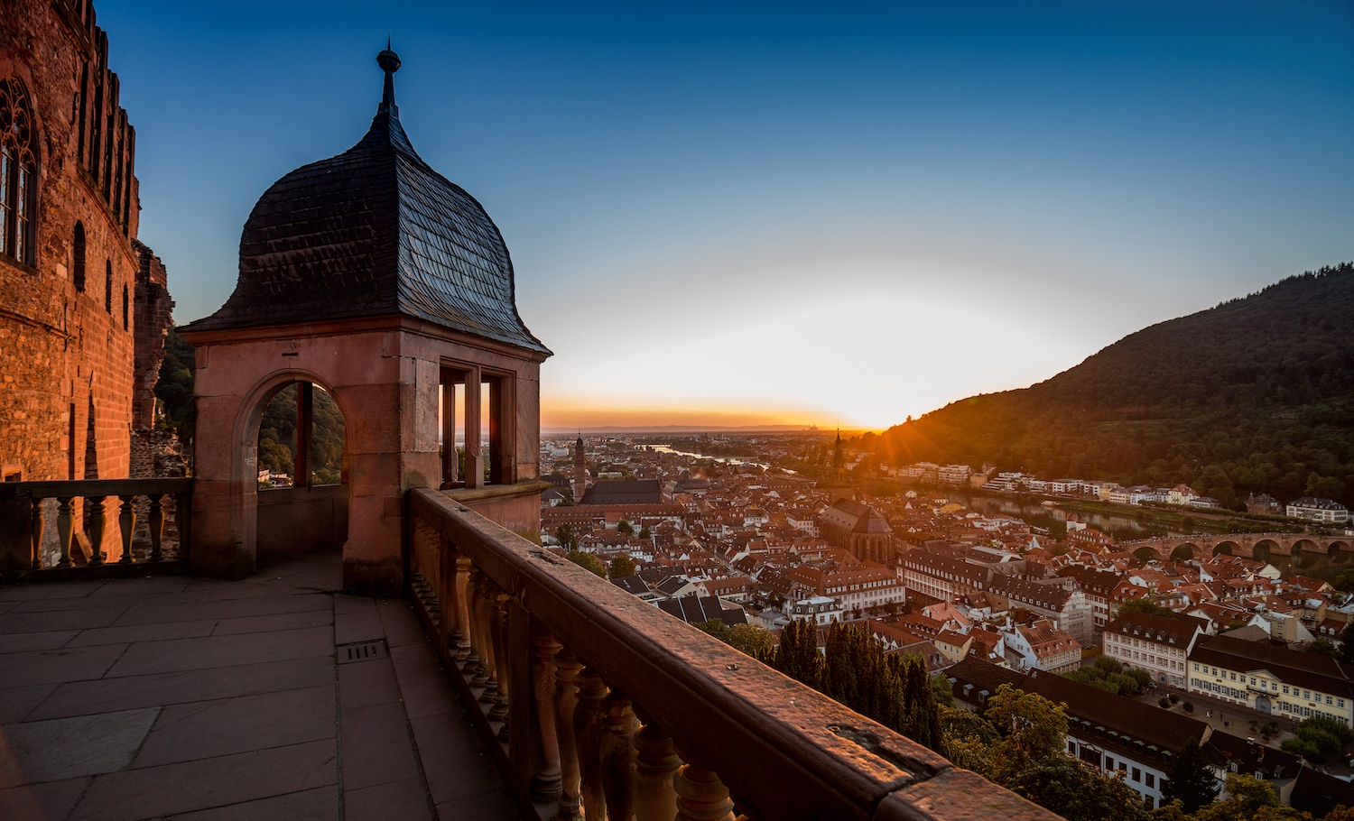 Blick von der Terrasse des Heidelberger Schlosses auf die Altstadt bei Sonnenuntergang. Im Vordergrund sind die historische Schlossmauer, eine Balustrade und ein Turm mit geschwungener Haube zu sehen. Im Hintergrund erstrecken sich die Dächer der Altstadt, der Neckar mit der Alten Brücke sowie bewaldete Hänge im warmen Abendlicht.