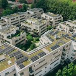 Luftaufnahme einer modernen Wohnanlage mit mehreren mehrgeschossigen Gebäuden, deren begrünte Flachdächer großflächig mit Photovoltaikanlagen ausgestattet sind. Die Häuser umschließen begrünte Innenhöfe und sind von altem Baumbestand umgeben. Das Bild veranschaulicht die Kombination aus urbanem Wohnungsbau, Dachbegrünung und dezentraler Solarstromerzeugung.