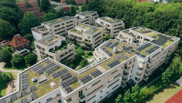 Luftaufnahme einer modernen Wohnanlage mit mehreren mehrgeschossigen Gebäuden, deren begrünte Flachdächer großflächig mit Photovoltaikanlagen ausgestattet sind. Die Häuser umschließen begrünte Innenhöfe und sind von altem Baumbestand umgeben. Das Bild veranschaulicht die Kombination aus urbanem Wohnungsbau, Dachbegrünung und dezentraler Solarstromerzeugung.