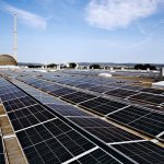 Blick über eine großflächige Photovoltaikanlage auf dem Flachdach eines Gewerbe- oder Industriegebäudes. Im Hintergrund sind ein markanter Schornstein, eine kuppelförmige Industrieanlage sowie weitere Gebäude und eine hügelige Landschaft unter blauem Himmel mit leichter Bewölkung zu sehen.