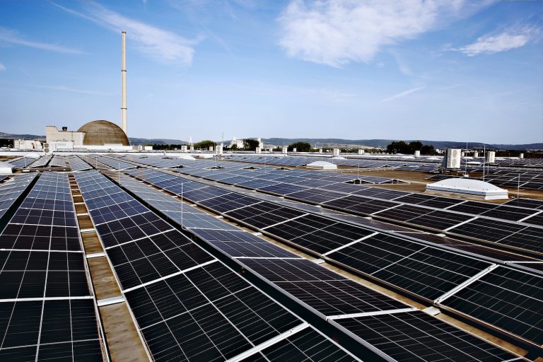 Blick über eine großflächige Photovoltaikanlage auf dem Flachdach eines Gewerbe- oder Industriegebäudes. Im Hintergrund sind ein markanter Schornstein, eine kuppelförmige Industrieanlage sowie weitere Gebäude und eine hügelige Landschaft unter blauem Himmel mit leichter Bewölkung zu sehen.