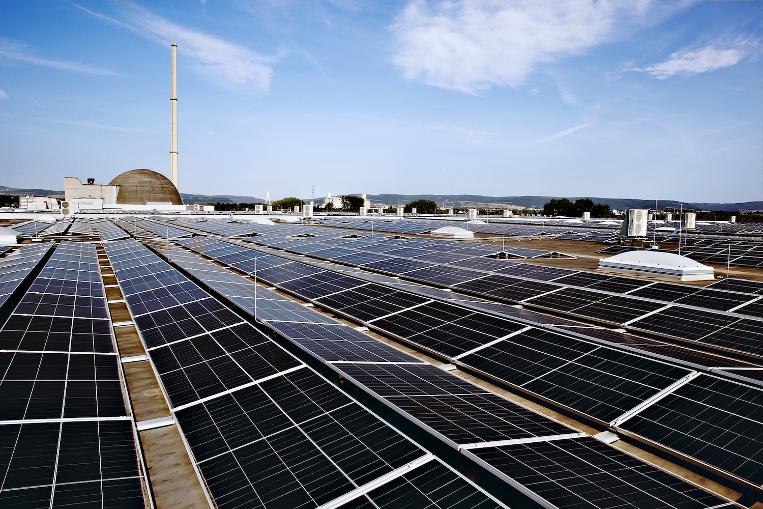 Blick über eine großflächige Photovoltaikanlage auf dem Flachdach eines Gewerbe- oder Industriegebäudes. Im Hintergrund sind ein markanter Schornstein, eine kuppelförmige Industrieanlage sowie weitere Gebäude und eine hügelige Landschaft unter blauem Himmel mit leichter Bewölkung zu sehen.