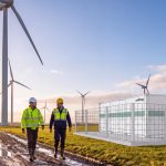 Zwei Mitarbeiter in Warnkleidung gehen auf einem schlammigen Weg durch einen Windpark. Rechts ist die Darstellung eines Batteriespeichers von Voltfang zu sehen.