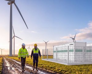 Zwei Mitarbeiter in Warnkleidung gehen auf einem schlammigen Weg durch einen Windpark. Rechts ist die Darstellung eines Batteriespeichers von Voltfang zu sehen.