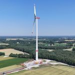 Luftaufnahme einer Windenergieanlage inmitten einer ländlichen Landschaft mit Feldern und Waldflächen. Im Hintergrund sind weitere Windräder zu sehen.