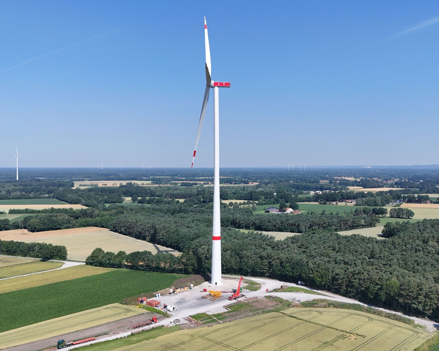 Luftaufnahme einer Windenergieanlage inmitten einer ländlichen Landschaft mit Feldern und Waldflächen. Im Hintergrund sind weitere Windräder zu sehen.