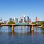 Blick über den Main auf die Frankfurter Skyline. Im Vordergrund überquert eine Straßenbahn auf einer Brücke den Fluss, dahinter erheben sich die Hochhäuser der Innenstadt und der Frankfurter Dom.
