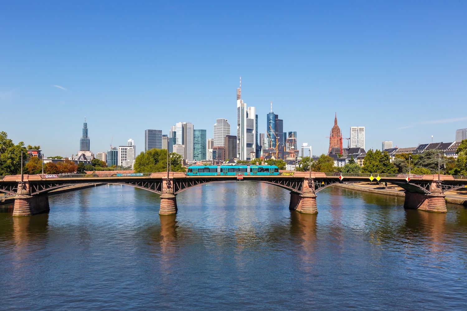 Blick über den Main auf die Frankfurter Skyline. Im Vordergrund überquert eine Straßenbahn auf einer Brücke den Fluss, dahinter erheben sich die Hochhäuser der Innenstadt und der Frankfurter Dom.