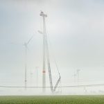 Mehrere Windenergieanlagen stehen im dichten Nebel. Im Vordergrund hebt ein großer Kran Bauteile für die Montage einer Windenergieanlage an.