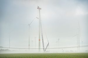 Mehrere Windenergieanlagen stehen im dichten Nebel. Im Vordergrund hebt ein großer Kran Bauteile für die Montage einer Windenergieanlage an.