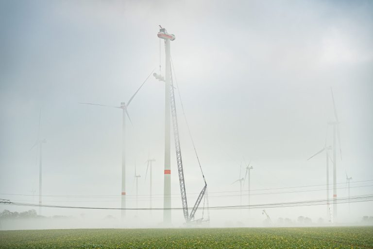 Mehrere Windenergieanlagen stehen im dichten Nebel. Im Vordergrund hebt ein großer Kran Bauteile für die Montage einer Windenergieanlage an.
