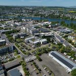 Luftaufnahme eines Gewerbegebiets am Moselbogen mit Büro- und Betriebsgebäuden, Parkplätzen und Photovoltaikanlagen auf mehreren Dächern. Im Hintergrund fließt die Mosel, dahinter erstrecken sich Wohngebiete, Felder und bewaldete Hügel.