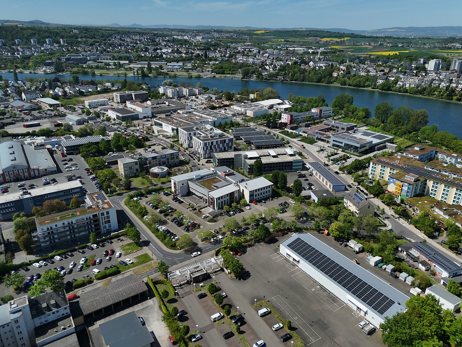 Luftaufnahme eines Gewerbegebiets am Moselbogen mit Büro- und Betriebsgebäuden, Parkplätzen und Photovoltaikanlagen auf mehreren Dächern. Im Hintergrund fließt die Mosel, dahinter erstrecken sich Wohngebiete, Felder und bewaldete Hügel.