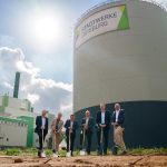 Sechs Projektbeteiligte beim symbolischen Spatenstich für die neuen Erzeugungsanlagen im Energiepark Wanheim der Stadtwerke Duisburg. Im Hintergrund sind Kraftwerksgebäude und ein großer Wärmespeicher zu sehen.