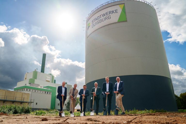 Sechs Projektbeteiligte beim symbolischen Spatenstich für die neuen Erzeugungsanlagen im Energiepark Wanheim der Stadtwerke Duisburg. Im Hintergrund sind Kraftwerksgebäude und ein großer Wärmespeicher zu sehen.