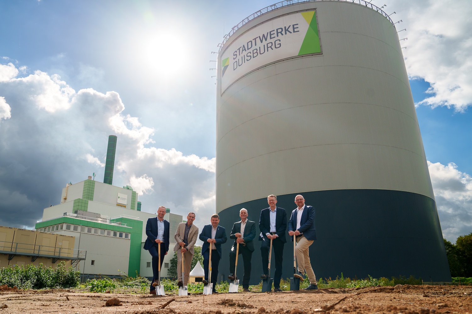 Sechs Projektbeteiligte beim symbolischen Spatenstich für die neuen Erzeugungsanlagen im Energiepark Wanheim der Stadtwerke Duisburg. Im Hintergrund sind Kraftwerksgebäude und ein großer Wärmespeicher zu sehen.