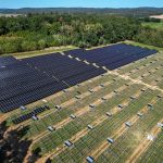 Luftaufnahme eines Solarparks in ländlicher Umgebung. Ein Teil der Photovoltaikmodule ist bereits installiert, während auf weiteren Flächen noch die Unterkonstruktionen zu sehen sind. Im Hintergrund erstrecken sich Wälder, Felder und Ortschaften.