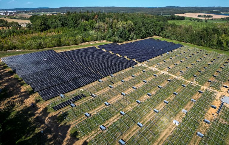 Luftaufnahme eines Solarparks in ländlicher Umgebung. Ein Teil der Photovoltaikmodule ist bereits installiert, während auf weiteren Flächen noch die Unterkonstruktionen zu sehen sind. Im Hintergrund erstrecken sich Wälder, Felder und Ortschaften.