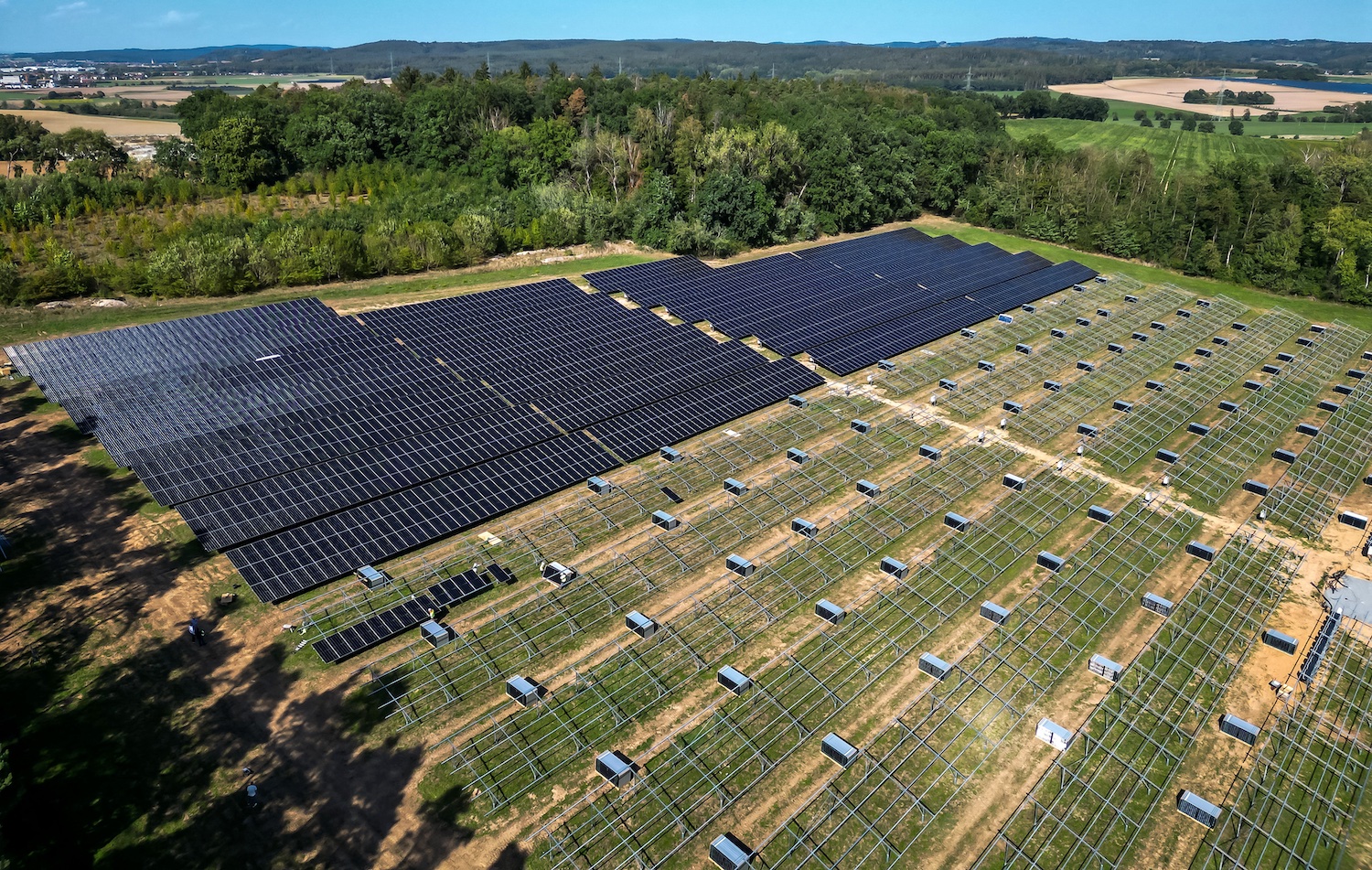 Luftaufnahme eines Solarparks in ländlicher Umgebung. Ein Teil der Photovoltaikmodule ist bereits installiert, während auf weiteren Flächen noch die Unterkonstruktionen zu sehen sind. Im Hintergrund erstrecken sich Wälder, Felder und Ortschaften.