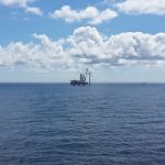 Blick auf einen im Bau befindlichen Offshore-Windpark auf dem Meer. In der Bildmitte steht ein Installationsschiff neben einer Windenergieanlage, weitere Anlagen sind am Horizont zu sehen.