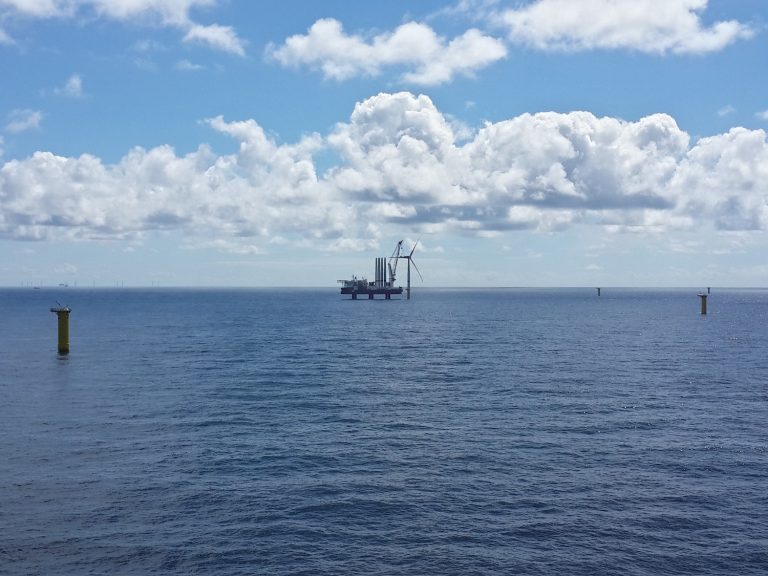 Blick auf einen im Bau befindlichen Offshore-Windpark auf dem Meer. In der Bildmitte steht ein Installationsschiff neben einer Windenergieanlage, weitere Anlagen sind am Horizont zu sehen.