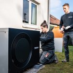Zwei Fachkräfte installieren die Außeneinheit einer Wärmepumpe von Buderus an der Fassade eines Wohnhauses.
